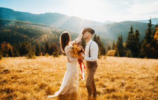 A-bride-in-a-white-dress-corrects-the-groom's-hat-at-sunset-as-they-take-rustic-wedding-photos-on-a-hillside.